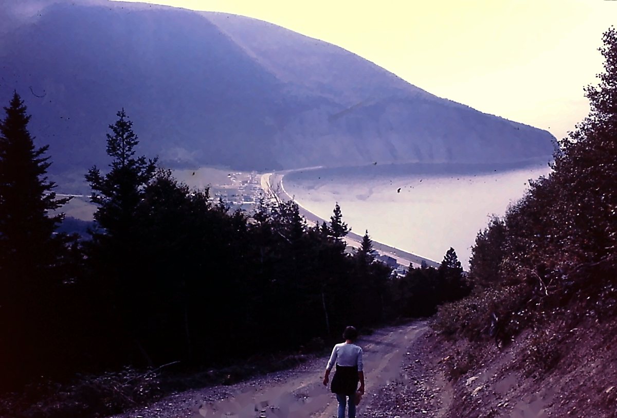 Ma mère, de dos, descend un chemin de gravier entre les épinettes ; en bas, le village, la longue plage en courbe et la baie, sous la grande montagne