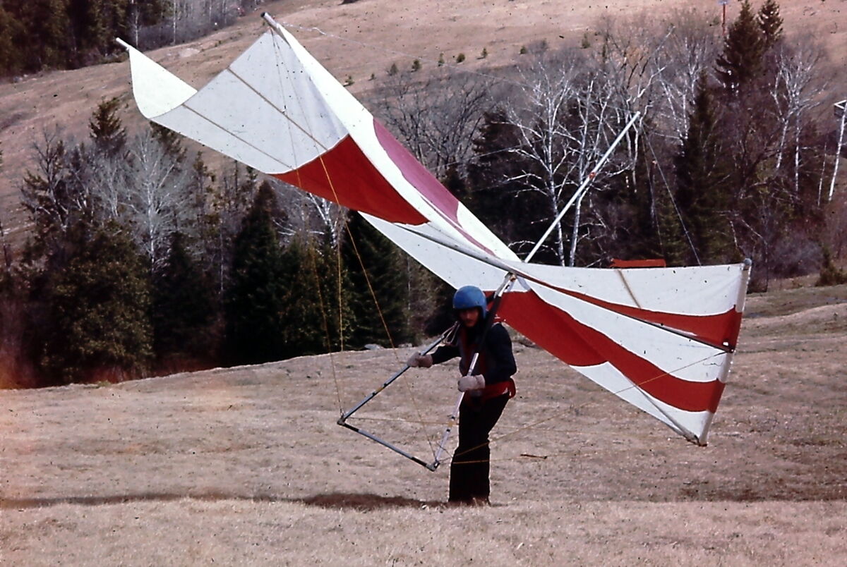 Un pilote au casque bleu porte un deltaplane rouge et blanc sur ses épaules, les mains sur la barre de contrôle, sur une pente d'herbe sèche bordée de conifères
