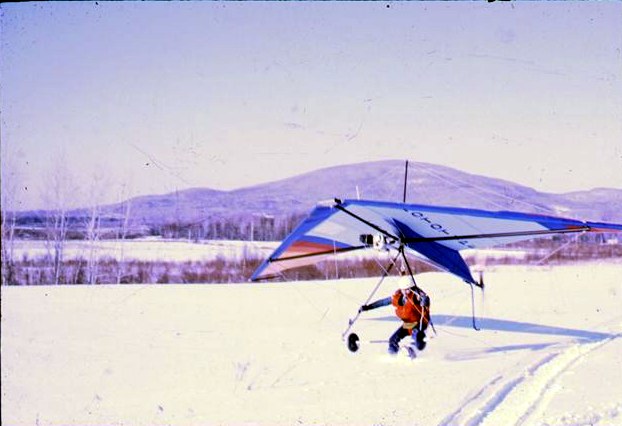 Une aile delta bleue et blanche munie de petites roues et d'un moteur sous la quille, le pilote debout dessous, dans un champ enneigé devant une montagne