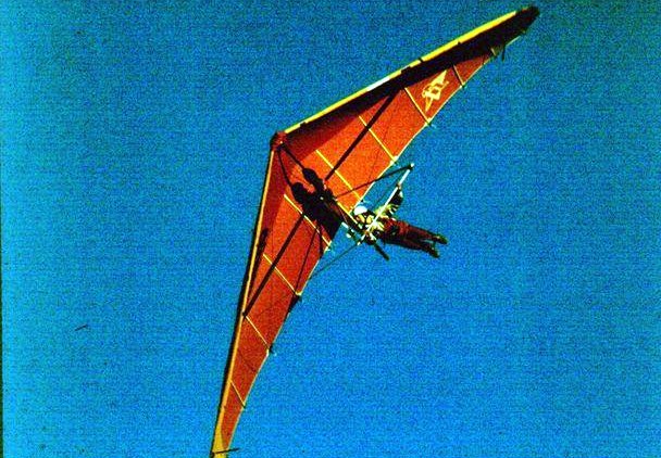 Une aile delta orange vue d'en dessous en plein vol, le pilote allongé sous la voile, sur un ciel bleu