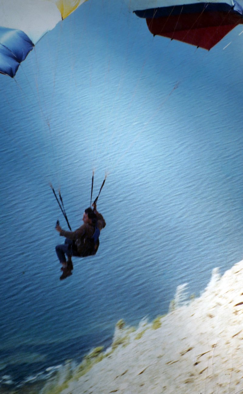 Un pilote assis dans sa sellette sous la voile du parapente, au ras de la pente du sommet, avec la mer loin en dessous