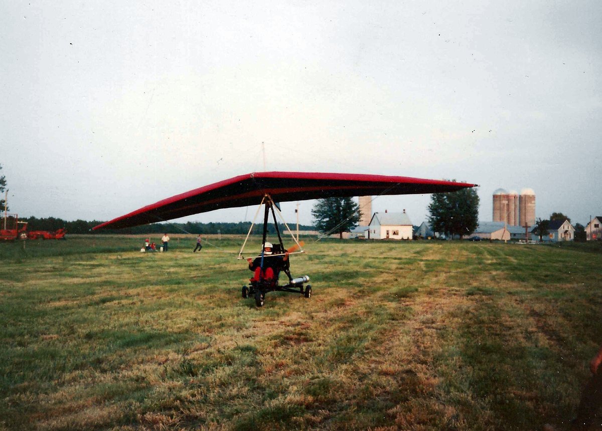 Un ULM à aile delta rouge sur un tricycle motorisé, le pilote casqué assis dessous, dans un champ fauché ; derrière, une maison blanche, des bâtiments de ferme et trois silos