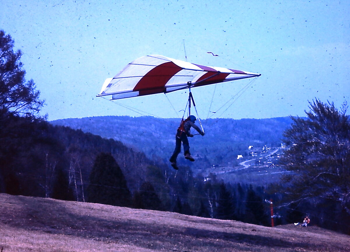 Un pilote casqué, suspendu sous un deltaplane rouge et blanc, vient de quitter la pente; derrière lui, une vallée boisée et un village
