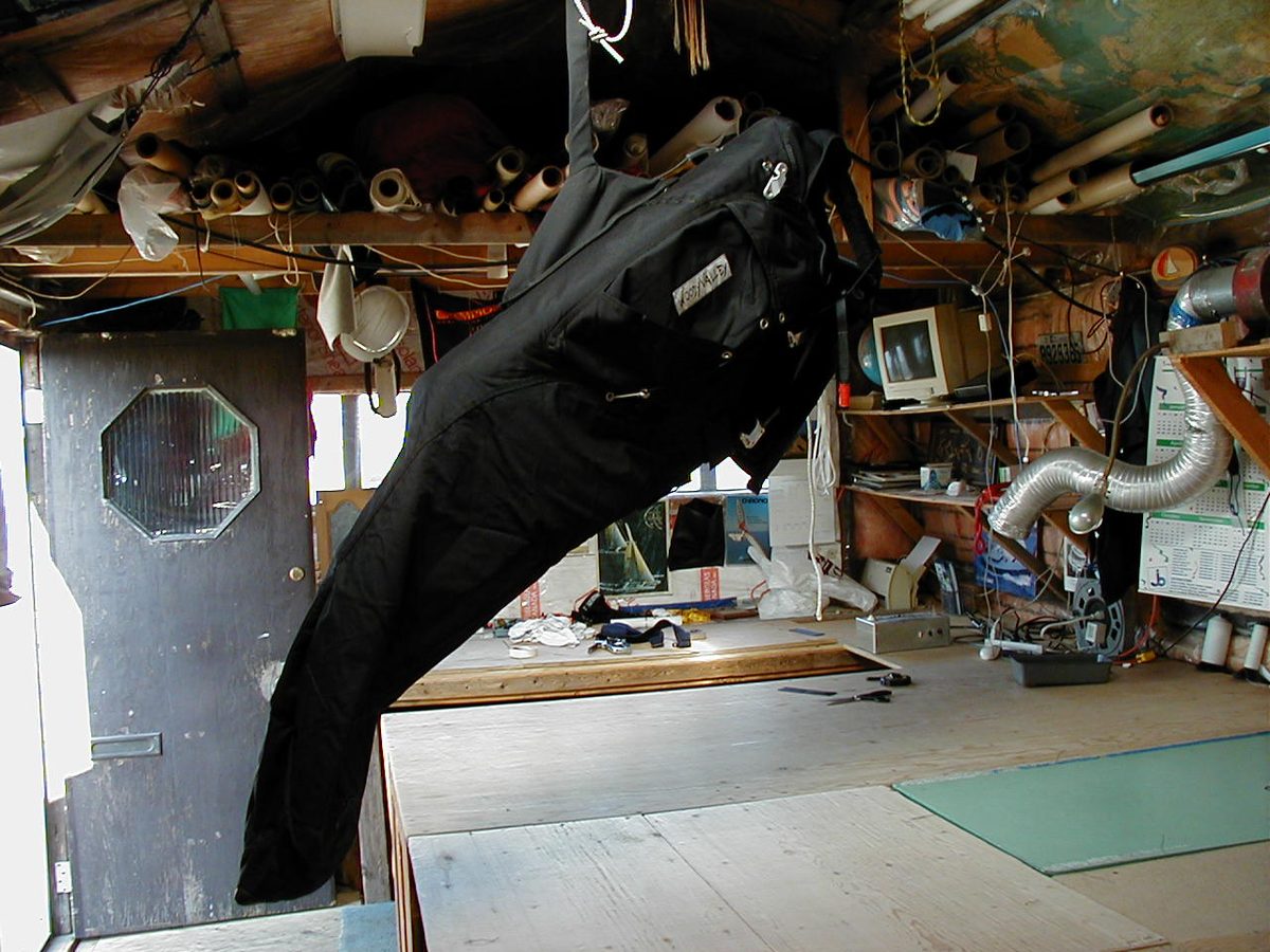 Un atelier dans un garage : des rouleaux de tissu rangés sous le plafond, une grande table de coupe, une porte à hublot octogonal, et un harnais de deltaplane noir suspendu au milieu