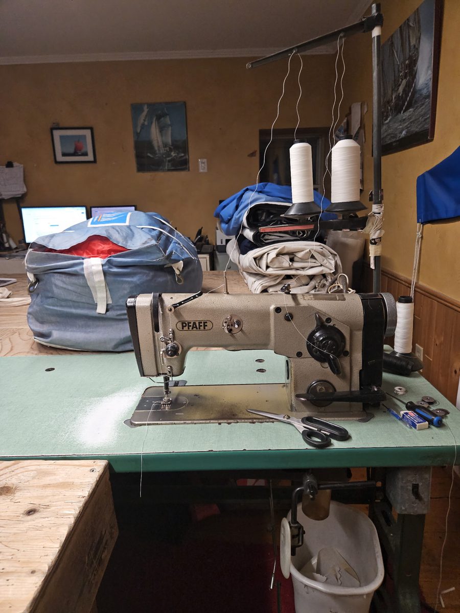 Une machine à coudre industrielle Pfaff, grise, sur une table verte, avec deux cônes de fil blanc sur son support, des voiles pliées et un sac de voile derrière