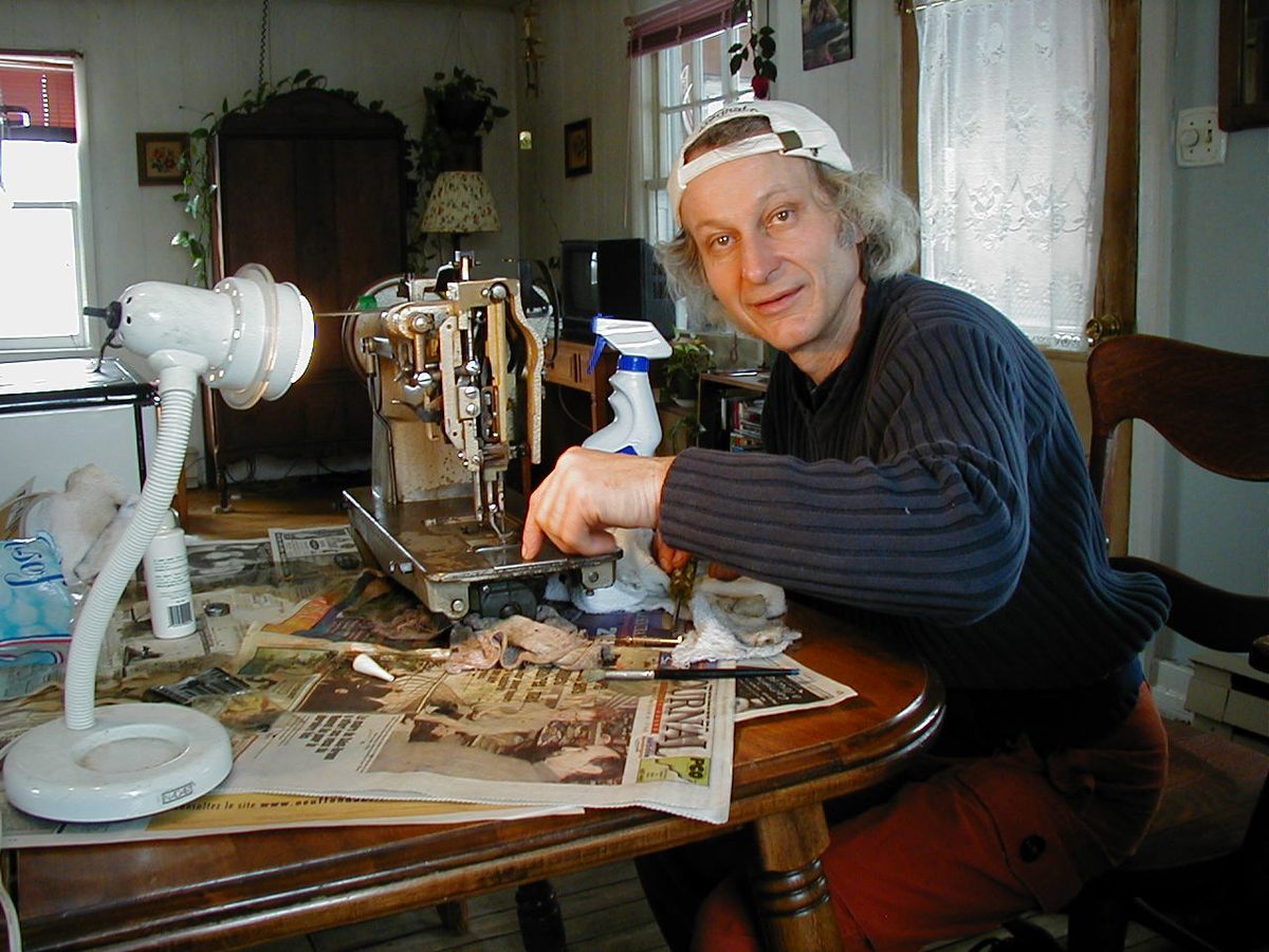 Un homme aux cheveux longs, casquette blanche à l'envers, répare une machine à coudre industrielle ouverte sur une table de salle à manger couverte de journaux, éclairée par une lampe de bureau