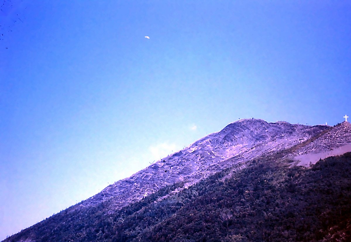Une montagne au sommet pelé sous un ciel bleu, une croix blanche sur la crête à droite; tout petit dans le ciel, un deltaplane