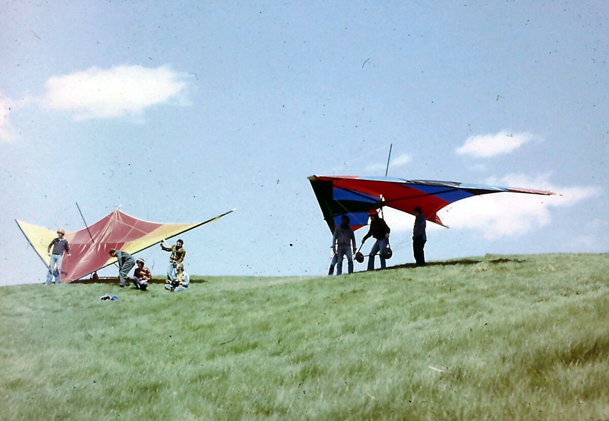 Le sommet gazonné d'une côte sous un ciel bleu; deux deltaplanes au sol, l'un jaune et rouge, l'autre rouge, bleu et noir, entourés d'une dizaine de personnes