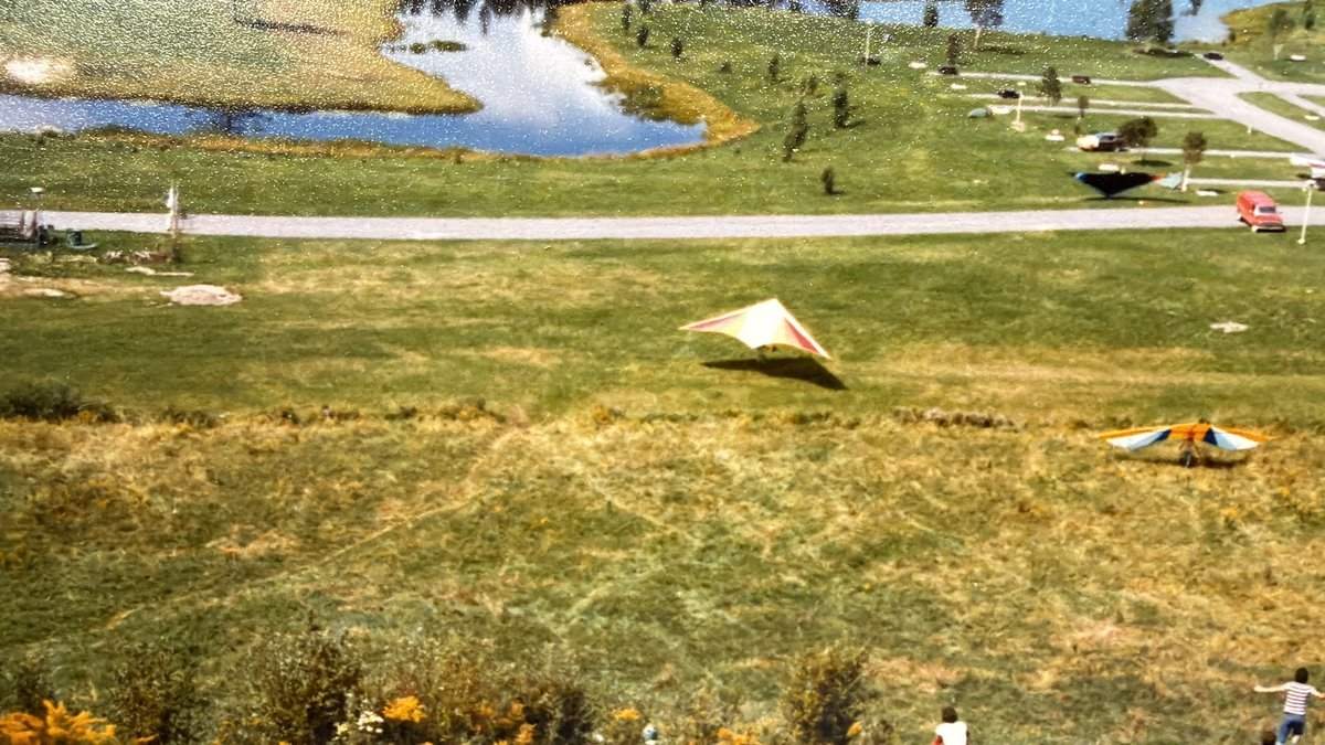 Vue du haut d'une pente herbeuse : un deltaplane pâle à bandes rouges glisse au ras du sol, un autre orange, bleu et blanc attend plus bas; au fond, un lac, les allées d'un camping et une fourgonnette rouge