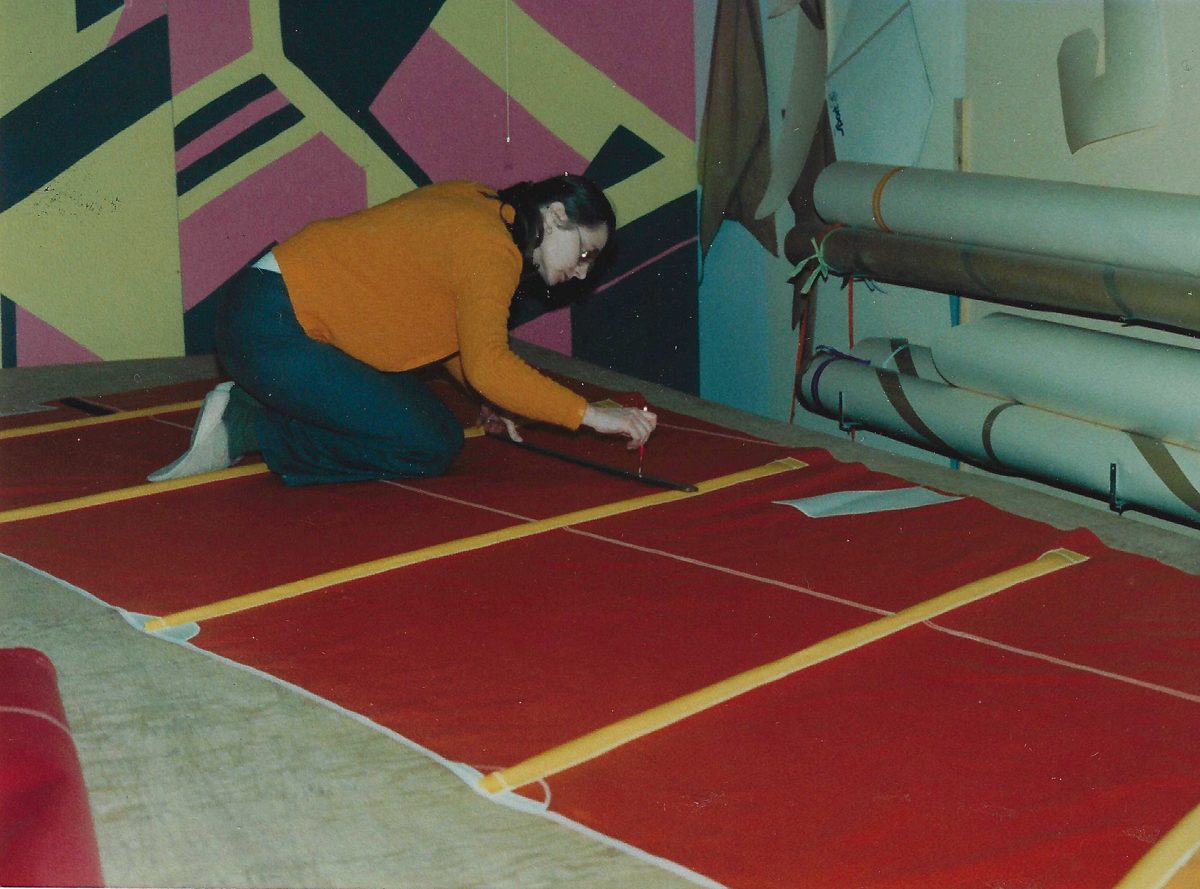 Une femme en chandail orange, à genoux sur un grand plancher couvert de tissu rouge, trace une ligne à la règle sur la voile ; derrière elle, un mur peint de grandes formes géométriques et des rouleaux de tissu accrochés au mur