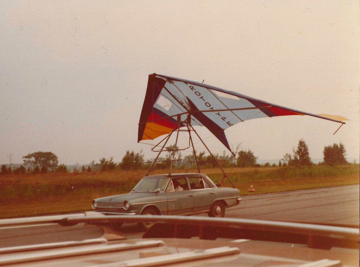 Une aile marquée PROTOTYPE, aux couleurs bleu, rouge, jaune et blanc, montée sur un bâti de tubes au-dessus d'une vieille berline grise, sur une piste d'aéroport bordée de champs