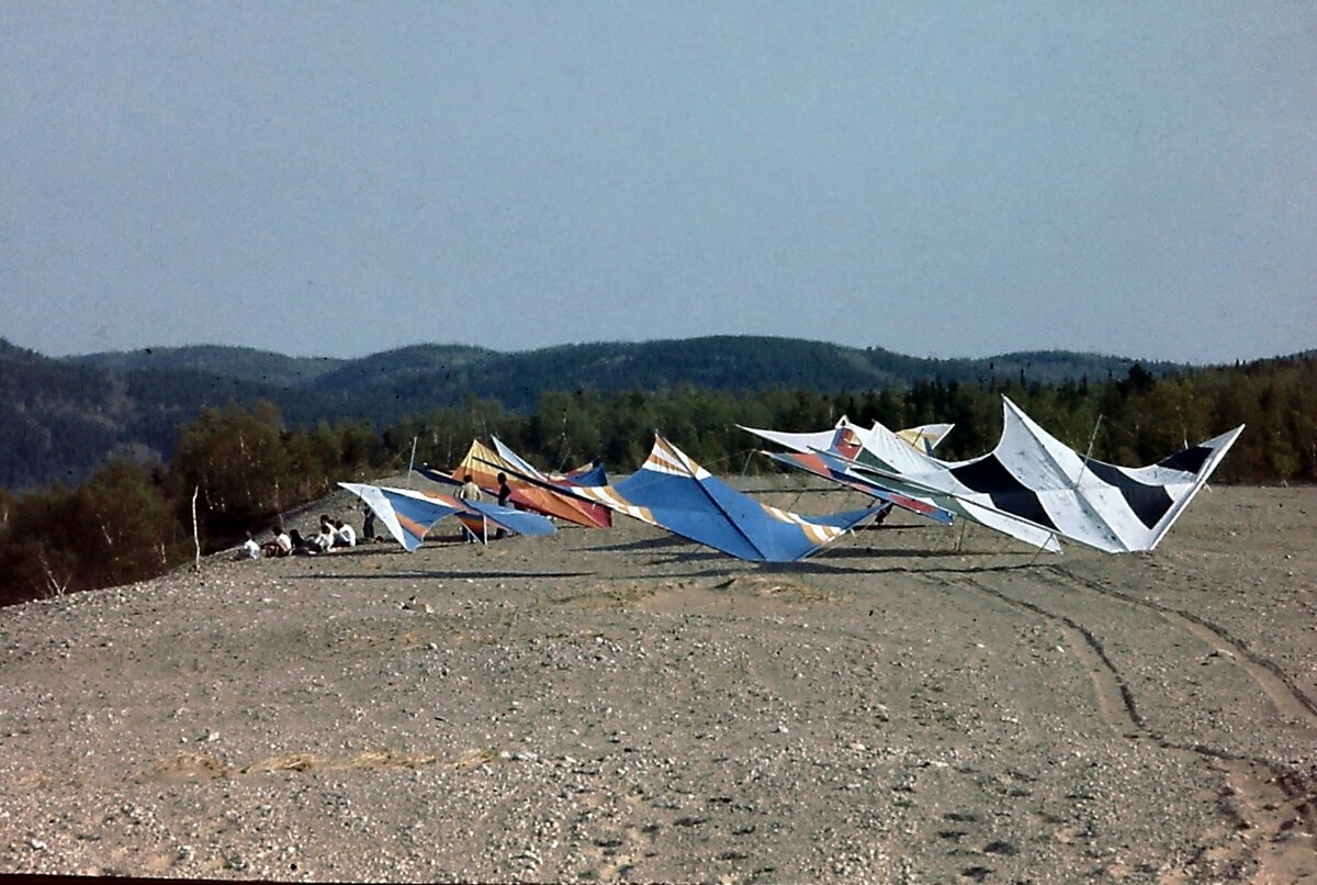 Une dizaine de deltaplanes de toutes les couleurs posés sur le sable d'une dune, devant des collines boisées; un groupe de pilotes est assis à gauche
