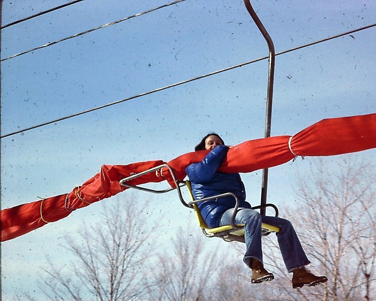 Une personne en manteau bleu assise sur un télésiège jaune serre dans ses bras un deltaplane plié dans sa longue housse orange; derrière, des arbres sans feuilles et le ciel d'hiver